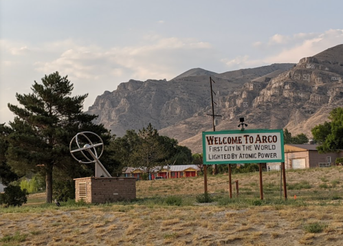 Sign welcoming visitors to Arco, Idaho, with mountains in the background and a sculpture nearby.