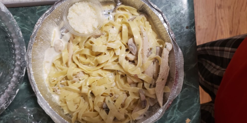 A serving of fettuccine alfredo with chicken and mushrooms, topped with grated cheese in a foil container.