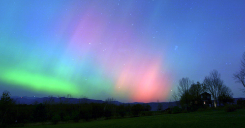 A vibrant aurora borealis lights up the night sky with green and pink hues over a landscape and distant house.
