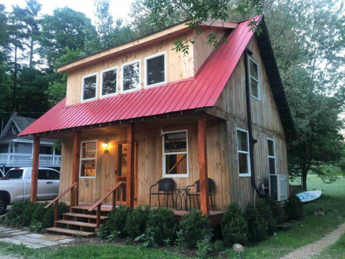 A cozy wooden cabin with a red metal roof, surrounded by greenery and a gravel path. Warm light glows from the windows.