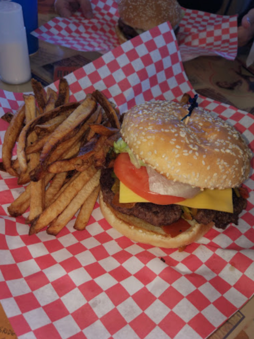 A sesame seed bun burger with cheese, lettuce, and tomato, served with a side of crispy French fries on a checkered paper.