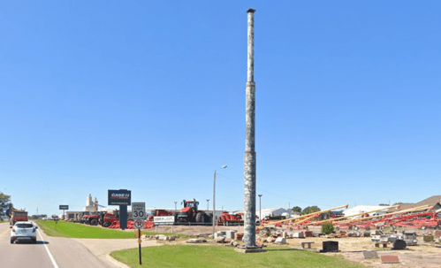 A tall, weathered pole stands beside a road, with farm equipment and a dealership sign in the background under a clear blue sky.