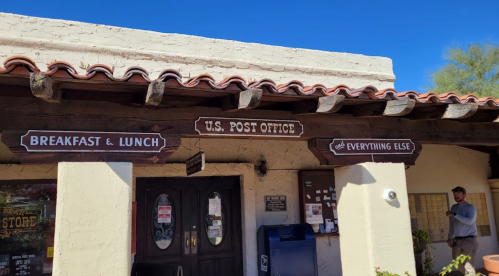A rustic building with signs for a U.S. Post Office, breakfast and lunch, and a general store. Clear blue sky above.