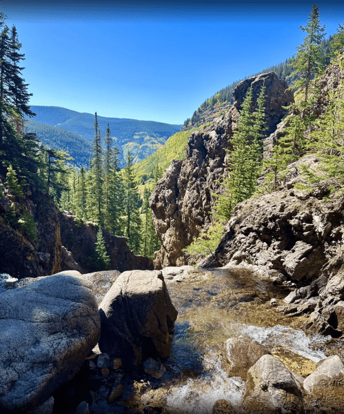 A serene landscape featuring a rocky stream surrounded by tall pine trees and mountains under a clear blue sky.