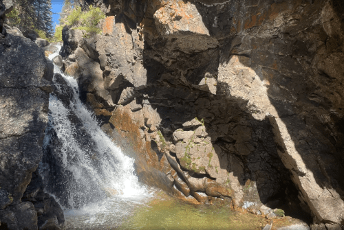 A small waterfall cascades over rocky cliffs into a serene pool below, surrounded by lush greenery.