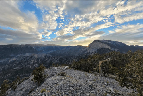 A panoramic view of mountains under a cloudy sky at sunset, with rocky terrain in the foreground.