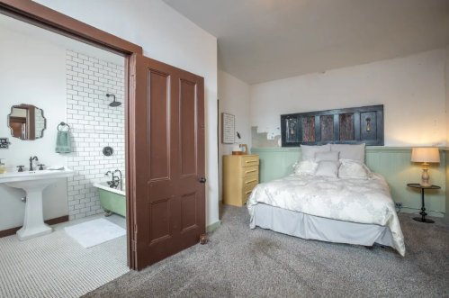 Cozy bedroom with a bed, dresser, and adjoining bathroom featuring a vintage tub and tiled wall.