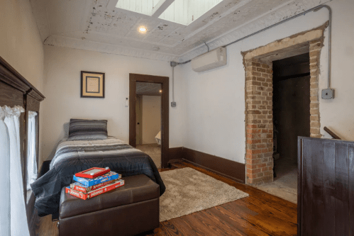 Cozy bedroom with a single bed, ottoman, and exposed brick wall, featuring a skylight and warm wooden floors.
