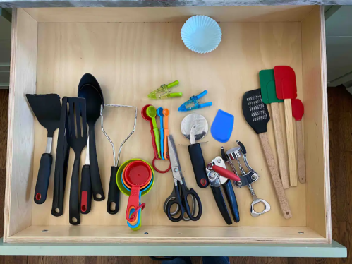 A wooden drawer filled with various kitchen utensils, including spatulas, measuring cups, scissors, and a corkscrew.