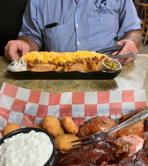 A man holds a large chili cheese dog, with a plate of fried food and dipping sauce in the foreground.