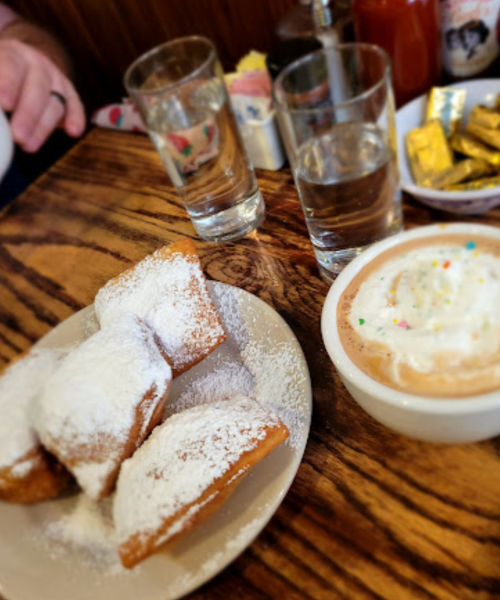 A plate of powdered sugar-dusted beignets next to a bowl of creamy dessert topped with whipped cream and sprinkles.