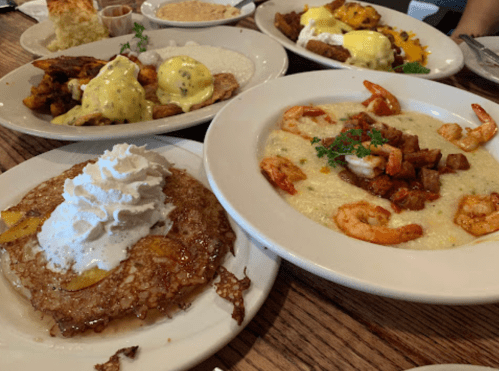 A table with various dishes: shrimp and grits, eggs Benedict, and a pancake topped with whipped cream and peaches.