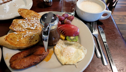 A plate of French toast, a fried egg, sausage, fresh fruit, and a cup of coffee on a wooden table.