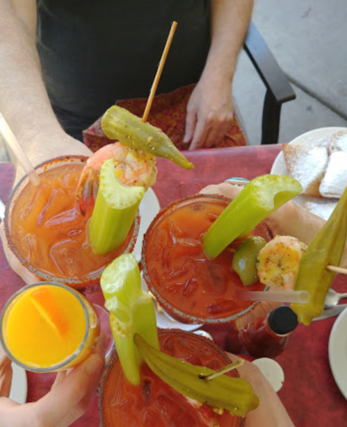 Four colorful cocktails with shrimp, celery, and pickles, served on a table with a small glass of orange juice.