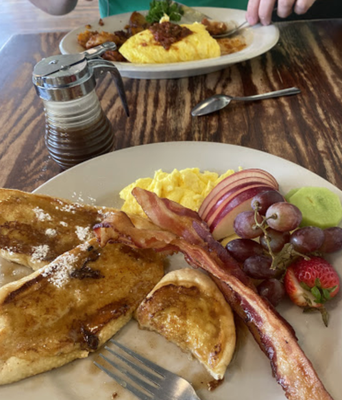 A plate of pancakes with syrup, scrambled eggs, bacon, and fruit, with another plate of omelet in the background.