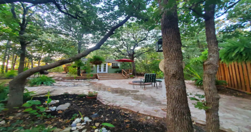 A serene backyard with stone pathways, greenery, and a cozy seating area near a small green building.