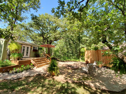 A sunny backyard with a wooden deck, patio furniture, and lush greenery under large trees.