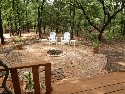 A circular brick patio surrounded by trees, featuring two white chairs and potted plants, with a fire pit in the center.