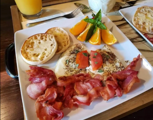 A plate with scrambled eggs, crispy bacon, English muffins, orange slices, and mint leaves, with a drink in the background.