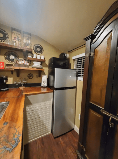 A cozy kitchen corner featuring a silver fridge, wooden countertop, and shelves with dishes and decor.