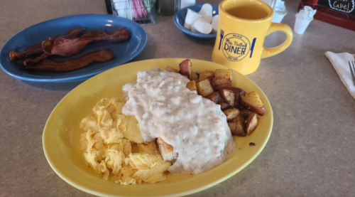 A plate of scrambled eggs, biscuits with gravy, and seasoned potatoes, with a side of bacon and a coffee mug.