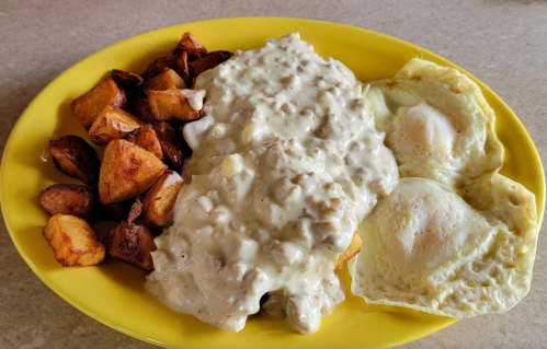 A yellow plate with two fried eggs, creamy sausage gravy over biscuits, and crispy potato cubes on the side.