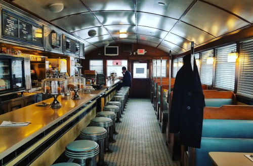 A vintage diner interior with a long counter, stools, and booths, featuring a man standing at the bar.