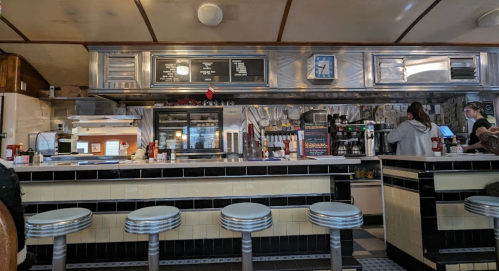 A retro diner interior featuring a counter with stools, a menu board, and staff preparing food in the background.