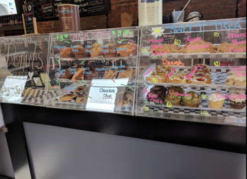 Display case filled with assorted donuts and pastries, labeled with flavors like chocolate chip, apple, and lemon.