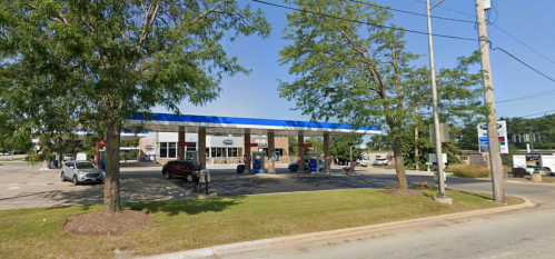 A gas station with a blue roof, surrounded by trees and parked cars, under a clear blue sky.