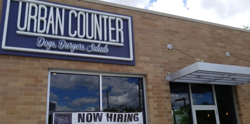 Exterior of Urban Counter restaurant with a "Now Hiring" banner displayed. The building features a modern design.