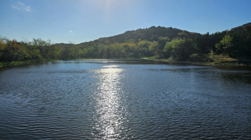 A serene river reflects sunlight, surrounded by lush greenery and hills under a clear blue sky.