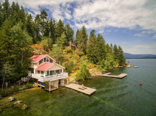 A scenic lakeside house with a red roof, surrounded by trees and docks on a calm water body under a blue sky.