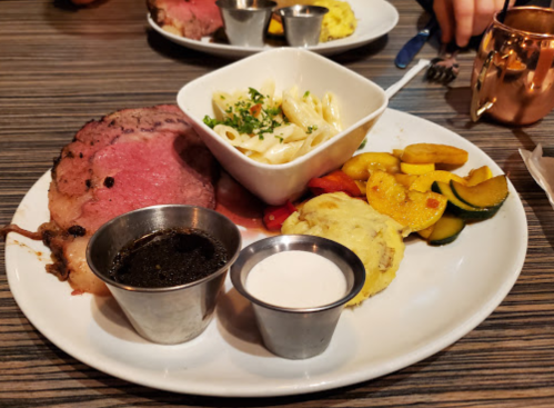 A plate with sliced roast beef, pasta salad, sautéed vegetables, and a small cup of sauce.