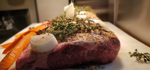 A raw beef roast seasoned with herbs and onions, accompanied by fresh carrots on a cutting board.