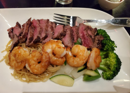 A plate of grilled steak slices, shrimp, broccoli, and noodles, garnished with vegetables.