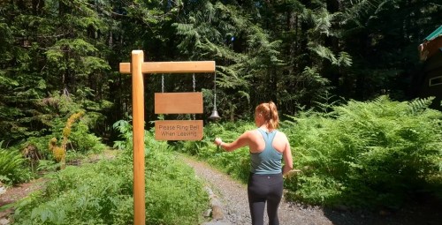 A person in activewear rings a bell at a trailhead sign surrounded by lush greenery and trees.