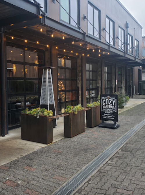 Exterior of a restaurant with string lights, planters, and a sign reading "Cozy Taverna" on a brick pathway.