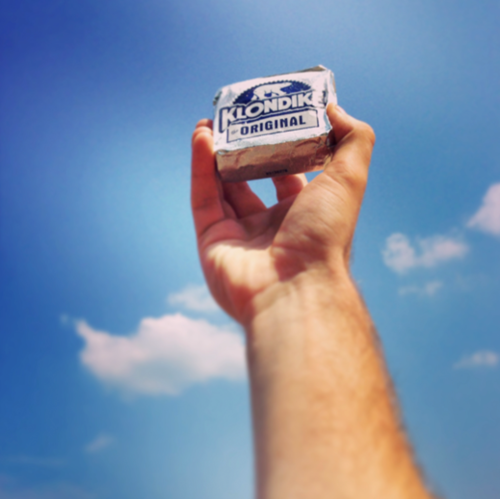 A hand holds a Klondike ice cream bar against a bright blue sky with fluffy clouds.