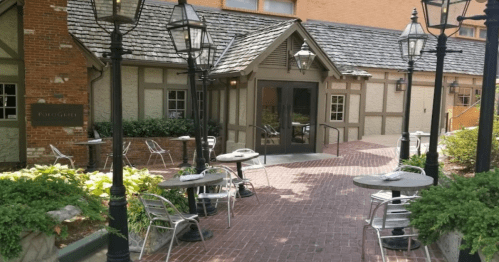 Outdoor dining area with tables and chairs, surrounded by greenery and lanterns, leading to a building entrance.