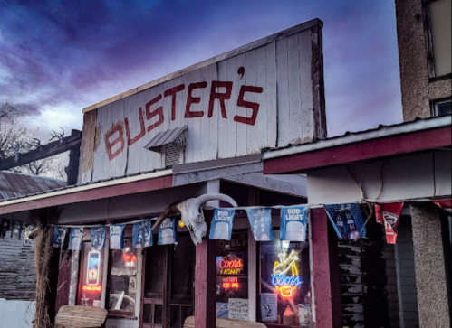 A rustic bar named "Buster's" with neon signs and banners, set against a colorful twilight sky.