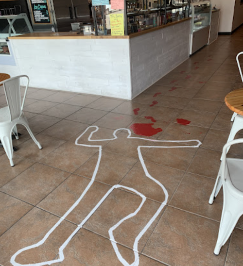 A chalk outline of a body on the floor with red stains, set in a café with a counter in the background.