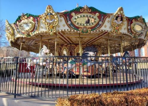 A colorful carousel with ornate decorations and horses, surrounded by a black fence and blue sky in the background.