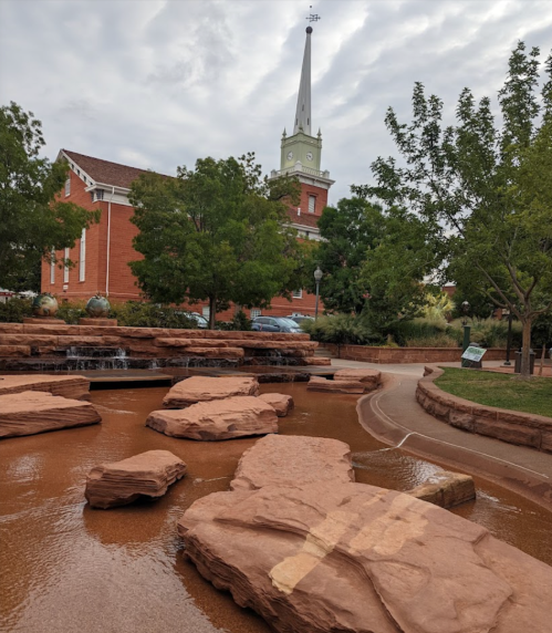 A serene park scene featuring large rocks, a water feature, and a church with a tall steeple in the background.