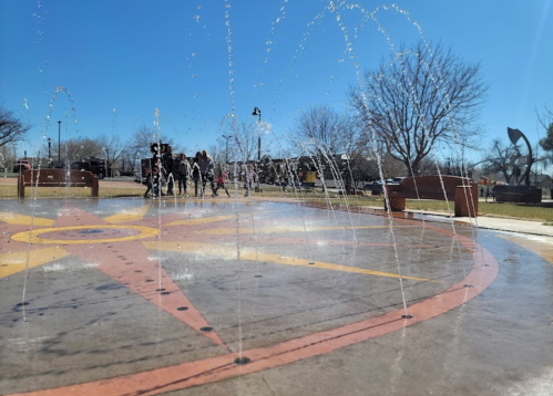 A sunny park scene featuring a colorful fountain with water jets, people walking in the background among trees.