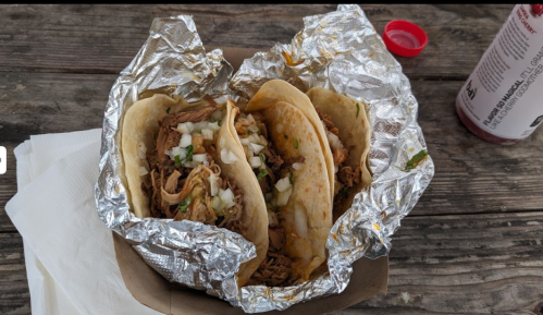 Three tacos filled with shredded meat, onions, and cilantro, wrapped in foil, on a wooden table.