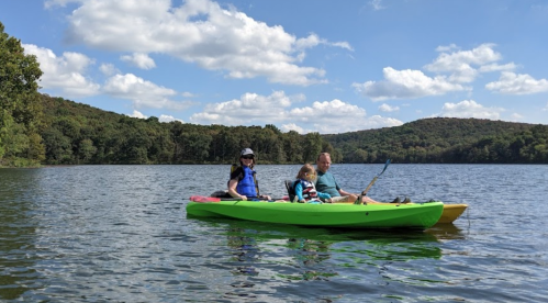 A family kayaking on a calm lake surrounded by green hills and blue skies with fluffy clouds.