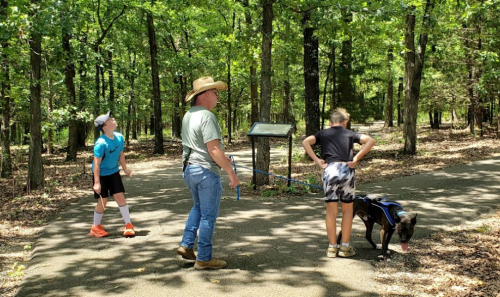 A man in a cowboy hat walks a dog on a leash, while two kids explore a wooded trail.