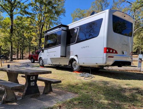 A sleek RV parked at a campsite, with a picnic table nearby and a red truck in the background under sunny trees.
