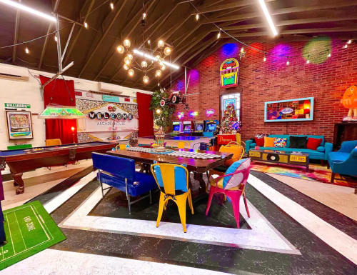 A vibrant game room with a pool table, arcade machines, colorful seating, and retro decor under warm lighting.